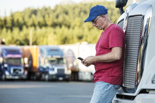 Caucasian man truck driver texting while standing next to the grill of his commercial truck in a struck stop.
